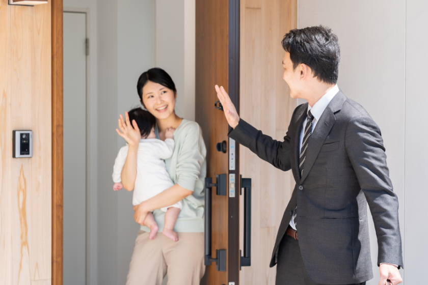 玄関で家族を見送るスーツ姿の男性と、子どもを抱く女性