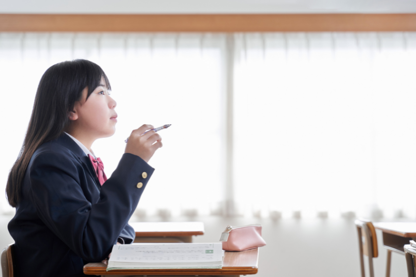 教室の窓辺でペンを持ちながら考える制服姿の女子生徒