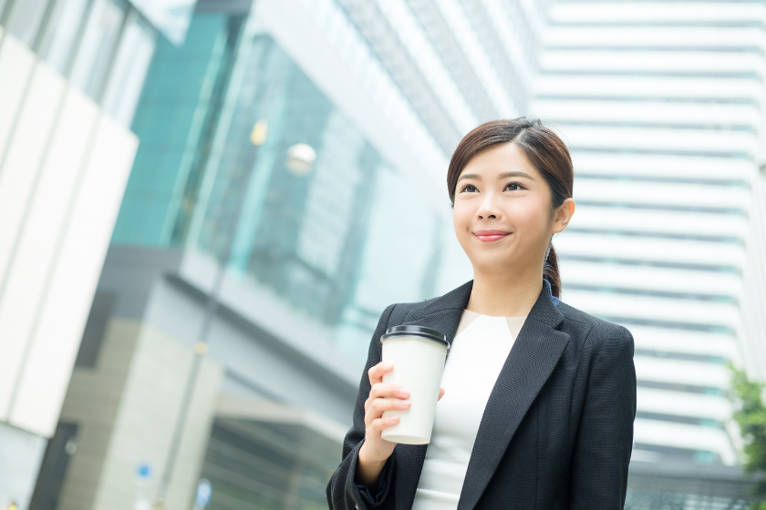 都市のビル街でカップを持ち、前を向くスーツ姿の女性