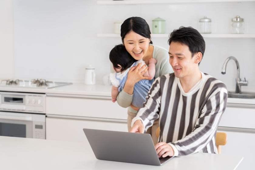 自宅でパソコン作業をする男性と、子どもを抱いて見守る女性